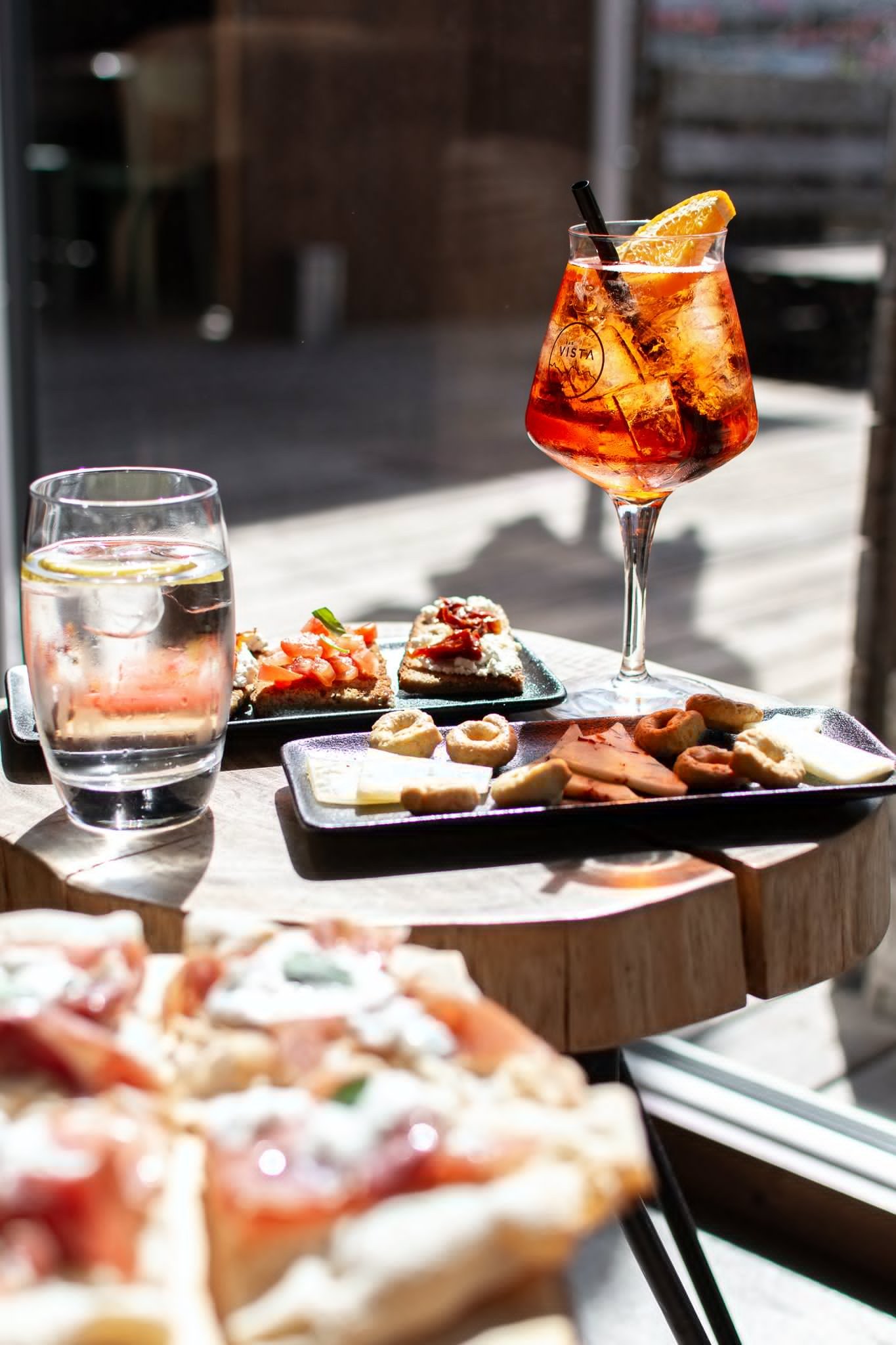 Aperitif, Wasserglas und kleine Speisen auf einem Holztisch der Vista Bar im Nachmittagslicht.