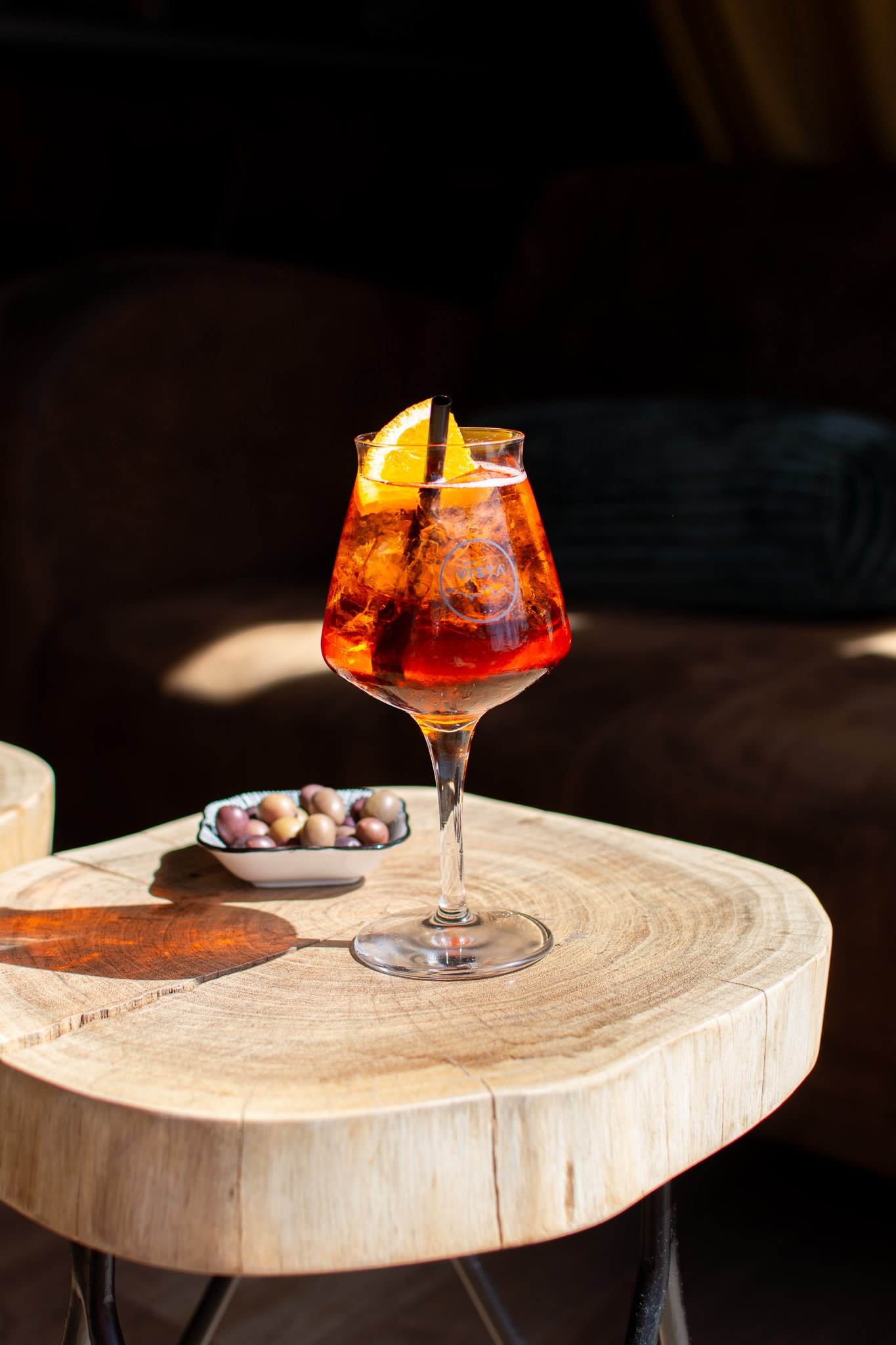Ein einzelnes Glas Aperitif auf einer Holztischplatte im warmen Abendlicht der Vista Bar.